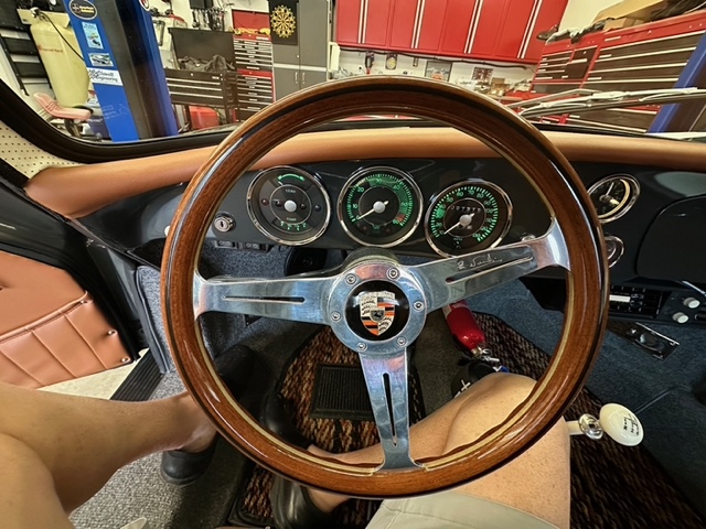 Wooden steering wheel and dashboard of a classic Porsche car, showing gauges and part of the driver’s legs.