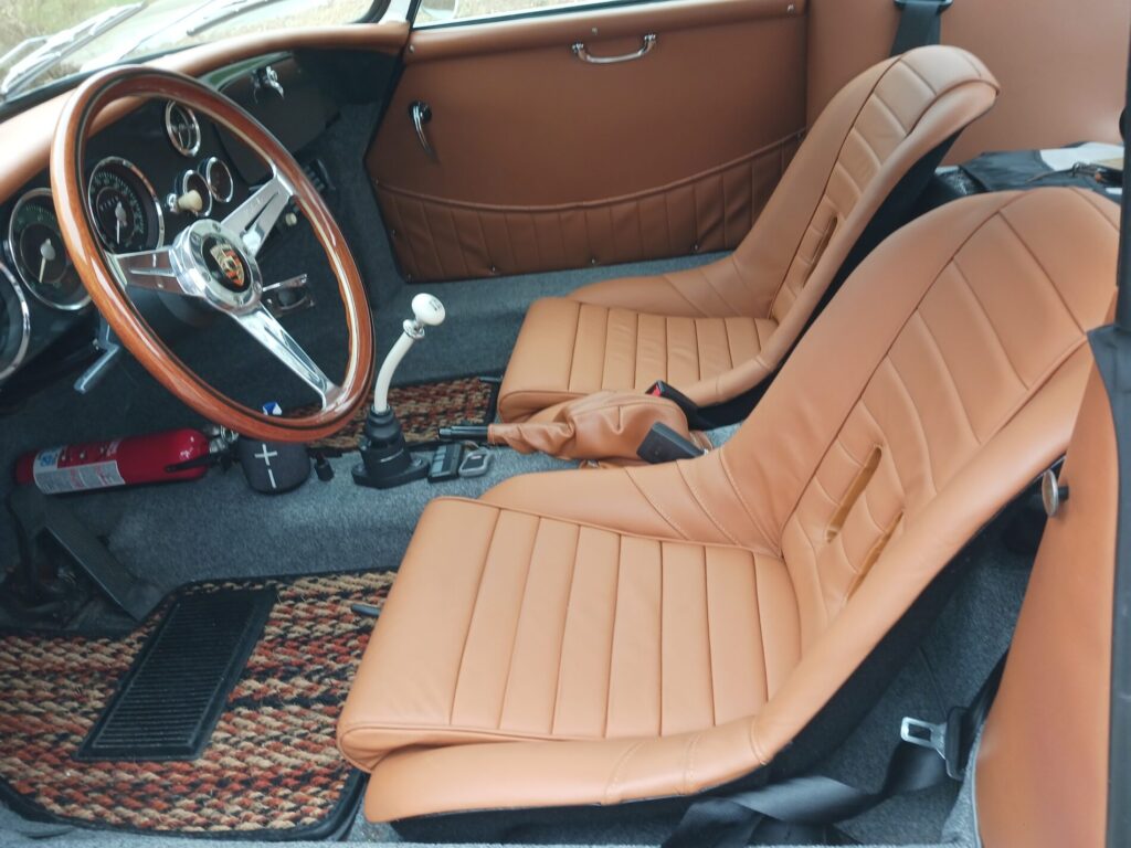 Vintage car interior with tan leather seats, wooden steering wheel, manual gear shift, and fire extinguisher by the door.