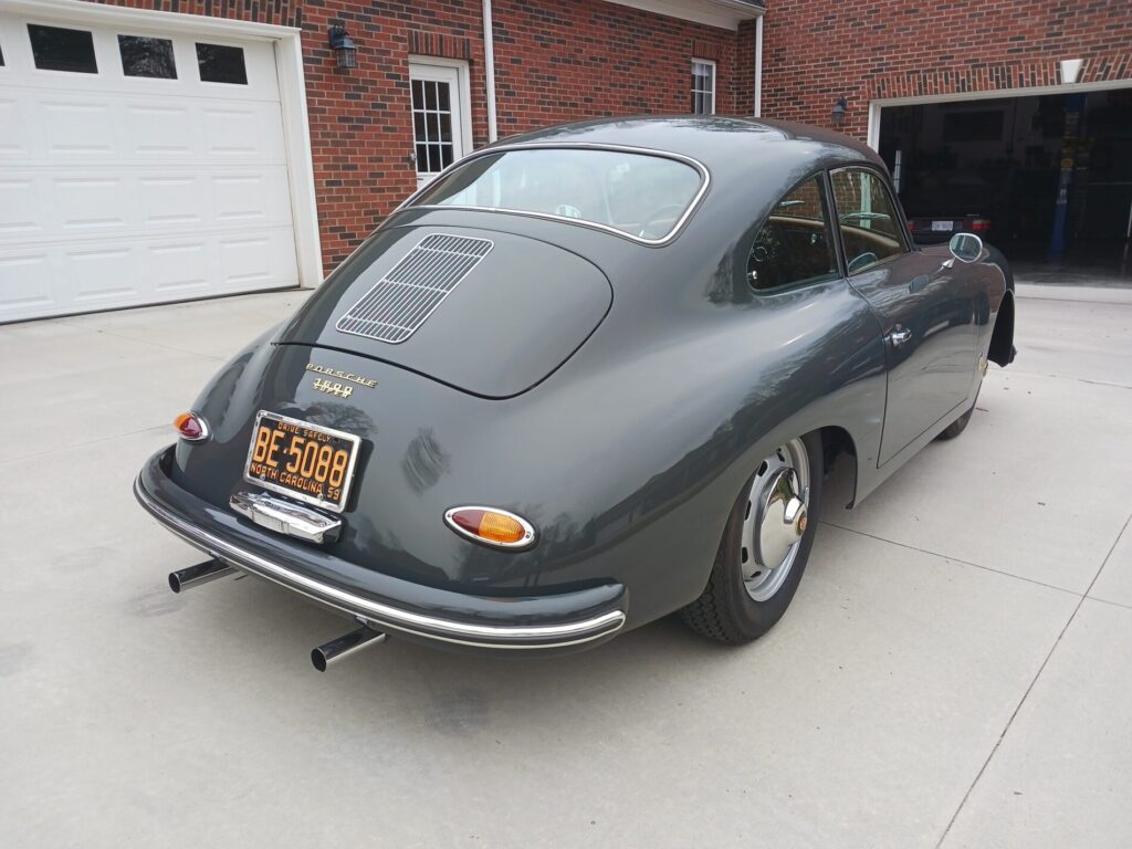 A classic gray Porsche 356A coupe is parked on a concrete driveway in front of a brick garage.