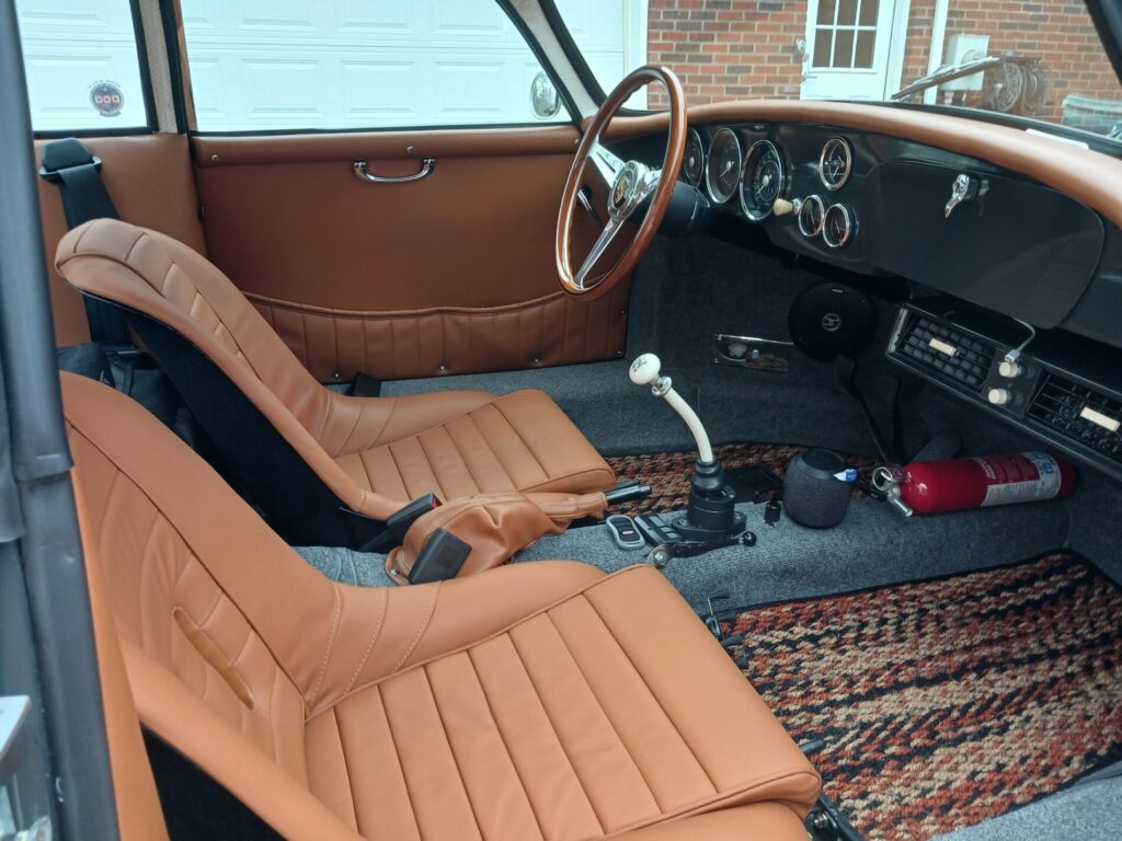 Vintage car interior with tan leather seats, classic steering wheel, manual gear shift, and a fire extinguisher on the floor.