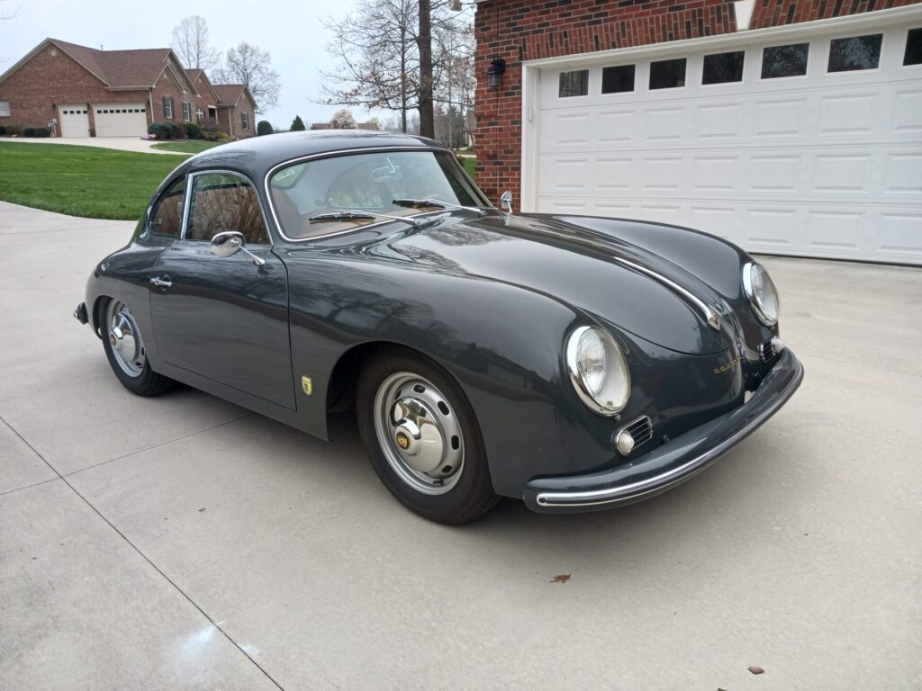A classic dark gray Porsche coupe parked on a concrete driveway in front of a brick house with a white garage door.