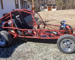 A red dune buggy with exposed frame and no body panels is parked on gravel near a house and a shed.