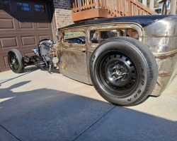 A customized, low-rider hot rod car with exposed metal body and black wheels parked in a driveway.