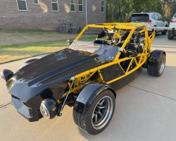 A black and yellow open-frame kit car is parked on a concrete driveway near a brick building and an SUV.