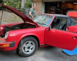 A red vintage car with its hood and door open is being inspected by two people in a driveway near a garage.