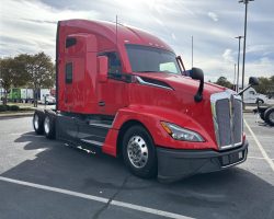 Red semi-truck parked in a lot on a sunny day, with trees and other vehicles in the background.